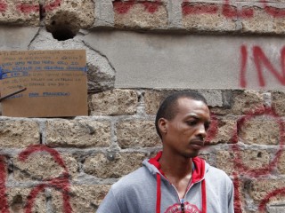 Josef, who said he arrived from Sudan 10 days earlier, holds clothes he received from volunteers as he stands on a street  in Rome July 14. In the background is a quote from Pope Francis about helping refugees. Several refugees said they were planning to head north to countries such as France and Germany. (CNS photo/Paul Haring) See VATICAN-MOBILE-MEDICAL July 14, 2016.