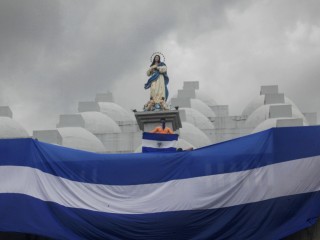 Một thanh niên đeo khẩu trang cầm quốc kỳ Nicaragua khi đứng trên nóc Nhà thờ Chính Tòa vào dịp lễ kỷ niệm một năm đánh dấu cuộc đàn áp của chính phủ đối với cuộc tuần hành vào Ngày của Mẹ ở Managua, Nicaragua, ngày 30 tháng 5 năm 2019. Cảnh sát Nicaragua đã cấm một cuộc cung nghinh Đức Mẹ đến Nhà thờ Chính Tòa dự kiến được tổ chức vào thứ Bảy, ngày 13 tháng 8 năm 2022 (Ảnh: Alfredo Zuniga / AP)